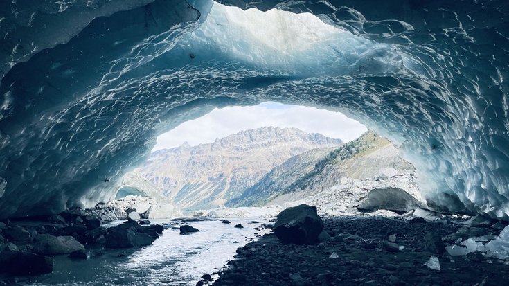 Blick aus einer Eishöhle, durch die ein kleiner Fluss fließt in eine alpine Landschaft