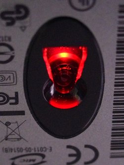 Detail-Ausschnitt der grauen Unterseite einer klassischen optischen Maus: In der Mitte zwischen diversen Logos und Sicherheitszeichen der Industrie sitzt ein dunkles Oval, in dessen Mitte eine kreisrunde, nach oben gewölbte Linse. Seitlich davon ein trapezförmiger, ebenfalls transparenter Ausschnitt, aus dessen Richtung rotes Licht leuchtet.