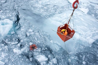 Ein Wissenschaftler steht in einer Gondel, die an einem Kran befestigt über einem mit Eisschollen durchsetzten Meeresabschnitt hängt. Mit einem langen weißen Stab will er ein orangefarbenes Messgerät aus dem Wasser bergen.