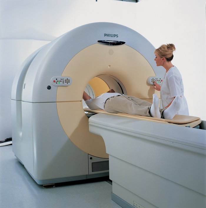 Patient wird von einer Schwester auf einer Liege in den Scanner geschoben. Der Scanner besteht aus einer großen Anlage mit zwei Ringen für CT und PET, die hintereinander angeordnet sind.