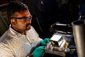 Ein Wissenschaftler mit Schutzbrille schließt in einem Labor Kabel an eine metallene Box an