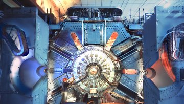 Eine große Maschine, in deren Mitte der ringförmige Beschleuniger zu erkennen ist. Als Größenvergleich steht ein Mensch neben dem Detektor, der etwa drei mal so groß wie der Mann ist.