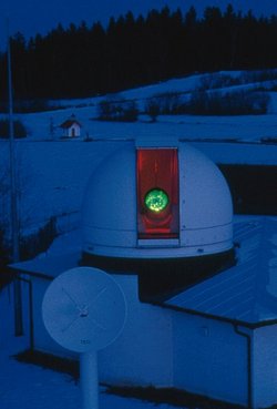 Nachtaufnahme in bläulichem Licht, geöffnete Kuppel mit grün leuchtendem Spiegel, im Vordergrund eine Parabolantenne