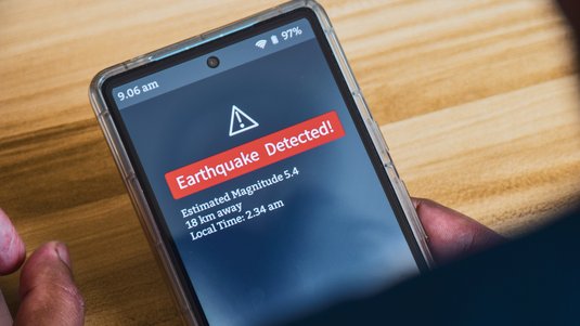 Eine Hand hält ein Smartphone, auf dem eine Erdbebenwarnung erscheint. Darauf steht: Earthquake Detected! Estimated Magnitude: 5,4. 18 km away. Local Time: 2:34 am.
