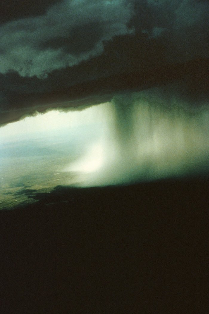 Foto. Aufnahme wurde von einem Flugzeug aus gemacht. Über einer Ebene befindet sich eine tiefdunkle Wolke, aus der ein grauer Schleier auf die Ebene hinab zu hängen scheint.