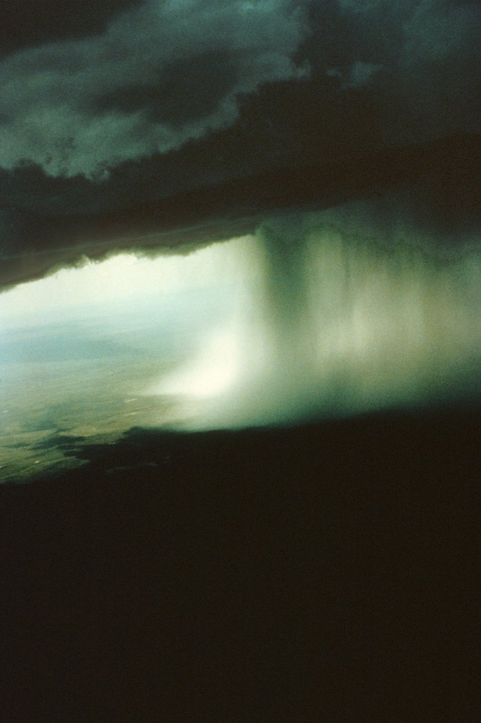 Luftaufnahme von Gewitterregen Foto. Aufnahme wurde von einem Flugzeug aus gemacht. Über einer Ebene befindet sich eine tiefdunkle Wolke, aus der ein grauer Schleier auf die Ebene hinab zu hängen scheint.
