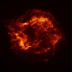 Eine runde, stark strukturierte Wolke aus hell- und dunkelroten Filamenten vor schwarzem Hintergrund; in der Mitte ein Stern.