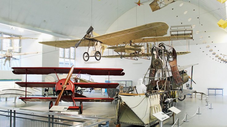 Mehrere einfache Flugzeugtypen - einige bestehen nur aus Sitz und segelartigen Flügeln - hängen von der Decke eines großen Museumsaals. Au dem Boden steht ein roter Dreidecker - also ein Flugzeug mit drei Tragflächen - und im Vordergrund eine schiffsa