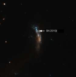 In der Mitte des Bildes eine längliche Struktur die blau und orange leuchtet. Vereinzelt sind Punkte zu erkennen. Ein kleiner Punkt ist mit einem weißen Pfeil mit SN 2010jl bezeichnet. Der Hintergrund ist schwarz mit vereinzelten, leuchtenden Punkten. 