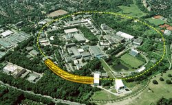 Luftfoto eines Forschungsgeländes, in das mit einer gestrichelten Linie ein kreisförmiger Beschleuniger eingezeichnet ist. Ebenfalls eingezeichnet ist eine große Halle, in die das Licht aus dem Beschleuniger geführt wird (durch gelbe Pfeile angedeutet).