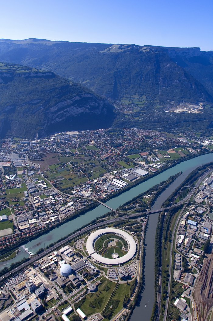 Luftaufnahme: Die ESRF ist ein helles kreisförmiges Gebäude, das sich in einer Stadt an der Mündung zweier Flüsse befindet.