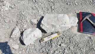 Zwei Gesteinsbrocken auf steinigem Boden. Einer von ihnen ist etwa so breit wie der danebenliegende Zollstock, der zweite nimmt ungefähr die gleich große Fläche wie das danebenliegende Buch ein.