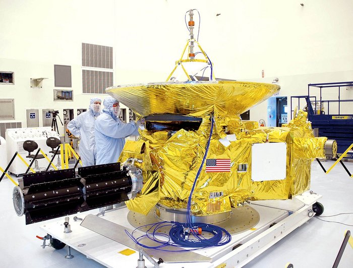 Eine in Goldfolie eingeschlagene Raumsonde in einem Reinraum. Zu erkennen ist sind große Isotopenbatterie und eine Satellitenschüssel. Menschen in Schutzanzügen arbeiten an der Sonde.