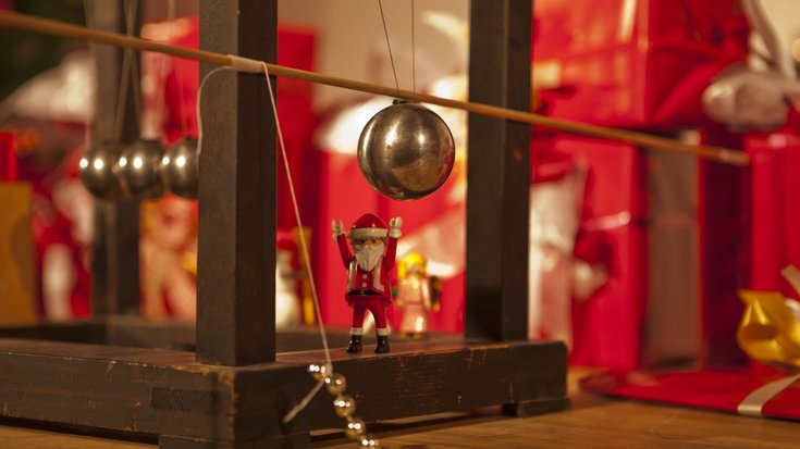 Weihnachtsmann als kleine Figur zwischen Geschenken und an Seilen aufgehängten Metallkugeln