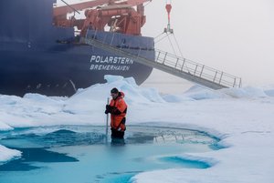 Mann in Schutzanzug steht in einem Schmelztümpel in einer Eislandschaft; im Hintergrund das Schiff Polarstern.