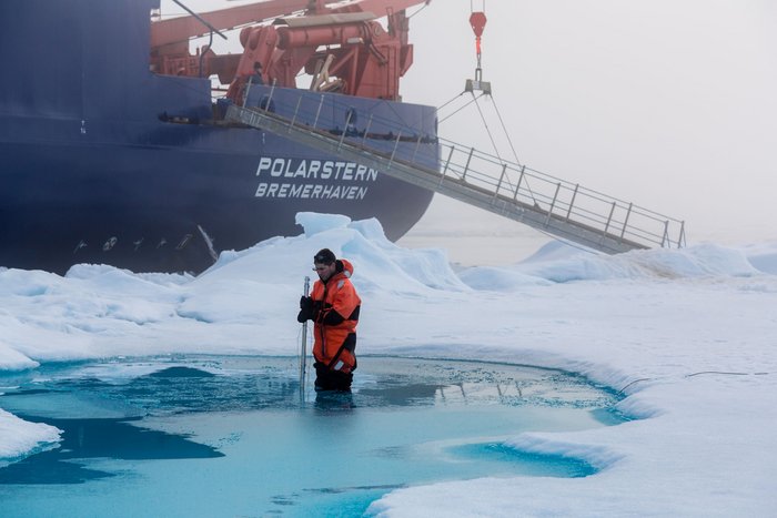 Marcel Nicolaus Mann in Schutzanzug steht in einem Schmelztümpel in einer Eislandschaft; im Hintergrund das Schiff Polarstern.
