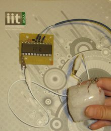 Eine Hand hält unten rechts einen hellen Plastikwürfel, von dem aus ein Kabel nach links oben führt, wo es mit einer Klemme an einer Platine mit Sensor befestigt ist. Von der Platine geht ein weiteres Kabel ab.