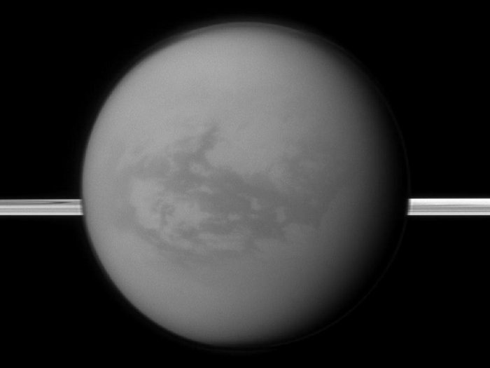 Schwarz-weiß-Aufnahme des Mondes Titan, auf der Oberfläche sind ein paar dunkle Strukturen zu erkennen, horizontal hinter dem Mond verlaufen die Saturnringe als heller Streifen.