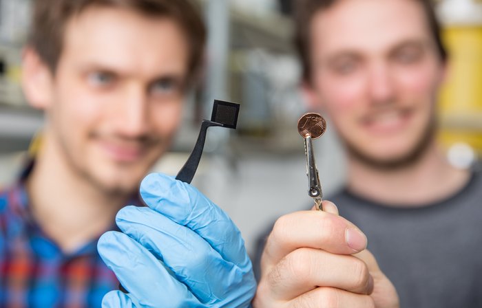 Ein Forscher hält den Mikrochip und ein anderer Forscher ein Ein-Cent-Stück. Beide sind etwa gleich groß.