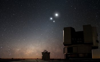 Im Hintergrund der Sternenhimmel. Der Mond, Jupiter und Venus sind deutlich als große helle Punkte zu sehen. Im Vordergrund zwei der vier Einzelteleskope, die zusammen das Very Large Telescope ergeben.  