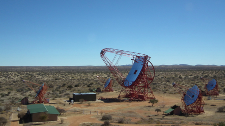 H.E.S.S II, Bildquelle: H.E.S.S. High Energy Stereoscopic System; Lizenz: gemäß den Bedingungen der Quelle Auf einer trockenen, braunen Landschaft mit einzelnen Büschen unter blauem Himmel steht im Zentrum des Bildes ein rostroter Aufbau mit einer kreisförmigen, dunklen, spiegelnden Oberfläche in der Mitte.