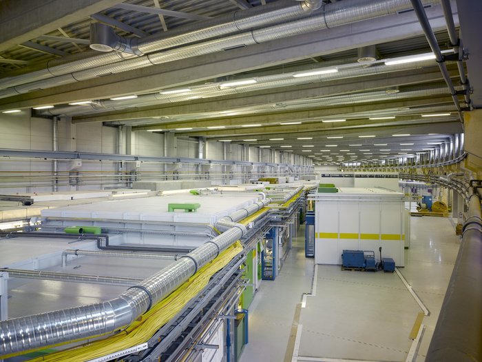 Blick in eine Halle, in der ein langes silbernes Rohr und Container mit Experimentierstationen zu sehen sind.