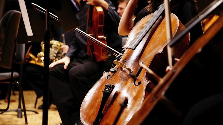 Ein Cello im Orchester. Im Hintergrund eine Geige und ein Horn. Die Musikerinnen und Musiker tragen dunkle Kleidung.