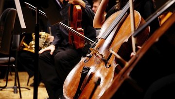 Ein Cello im Orchester. Im Hintergrund eine Geige und ein Horn. Die Musikerinnen und Musiker tragen dunkle Kleidung.
