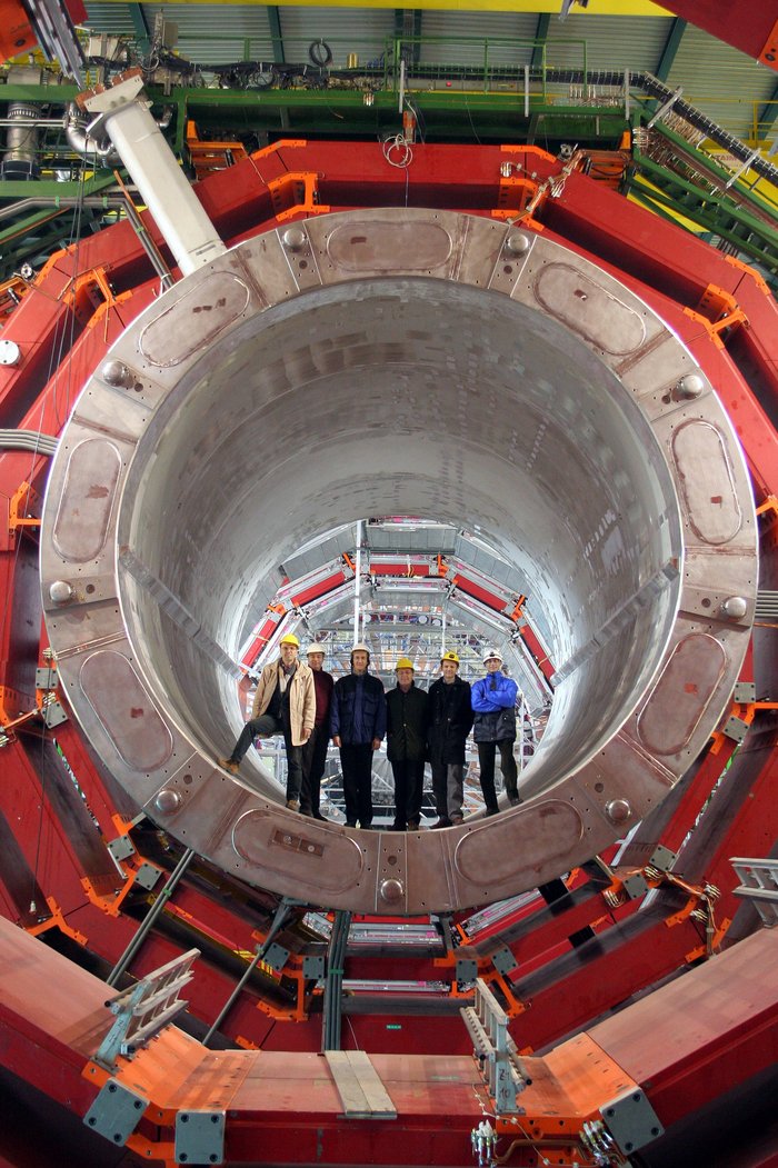 Sechs Personen stehen einem großen grauen Zylinder, der von einer roten Stahlstruktur umgeben ist.