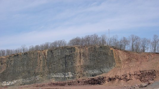 Foto einer steilen rötlich-schwarzen Gesteinswand unter blauem Himmel, an der Sedimentstreifen sichtbar sind.