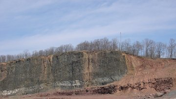 Foto einer steilen rötlich-schwarzen Gesteinswand unter blauem Himmel, an der Sedimentstreifen sichtbar sind.