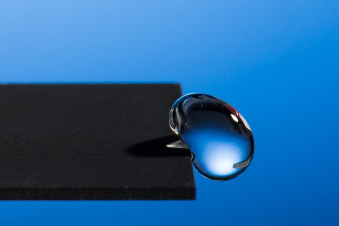 Abperlender Wassertropfen Ein Wassertropfen an einem schwarzen Material auf blauem Hintergrund.