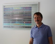 Thomas Henning steht vor einem Poster, das Ansichten des Milchstraßensystems in verschiedenen Wellenlängen zeigt. 