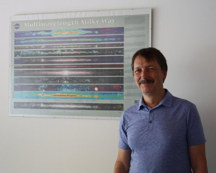Thomas Henning vom Max-Planck-Institut für Astronomie in Heidelberg Thomas Henning steht vor einem Poster, das Ansichten des Milchstraßensystems in verschiedenen Wellenlängen zeigt.