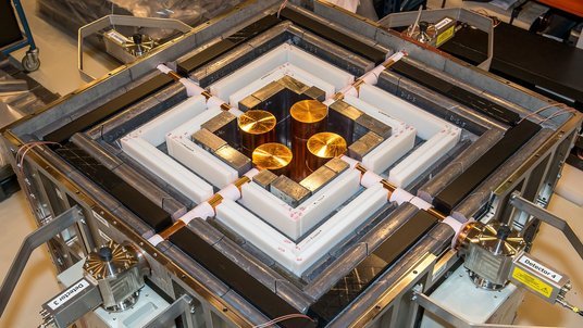 Quadratische technische Vorrichtung, die durch verschiedene Rahmen umgeben ist; in der Mitte befinden sich vier goldene Scheiben, die durch Kabeln mit dem Äußeren der Maschine verbunden sind
