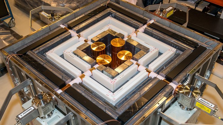 Quadratische technische Vorrichtung, die durch verschiedene Rahmen umgeben ist; in der Mitte befinden sich vier goldene Scheiben, die durch Kabeln mit dem Äußeren der Maschine verbunden sind