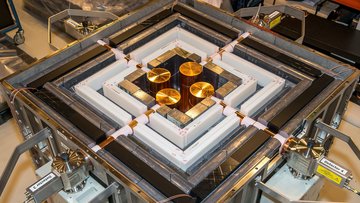 Quadratische technische Vorrichtung, die durch verschiedene Rahmen umgeben ist; in der Mitte befinden sich vier goldene Scheiben, die durch Kabeln mit dem Äußeren der Maschine verbunden sind