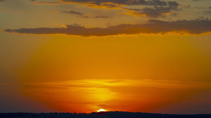 Sonnenuntergang über einer dunklen Hügelsilhouette. Die glutrote Sonne steht knapp über dem Horizont, der Himmel leuchtet von rot über orange bis gelb und wird nach oben hin bläulich, durchzogen von wenigen Wolkenbändern.
