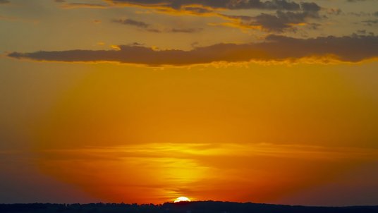 Sonnenuntergang über einer dunklen Hügelsilhouette. Die glutrote Sonne steht knapp über dem Horizont, der Himmel leuchtet von rot über orange bis gelb und wird nach oben hin bläulich, durchzogen von wenigen Wolkenbändern.