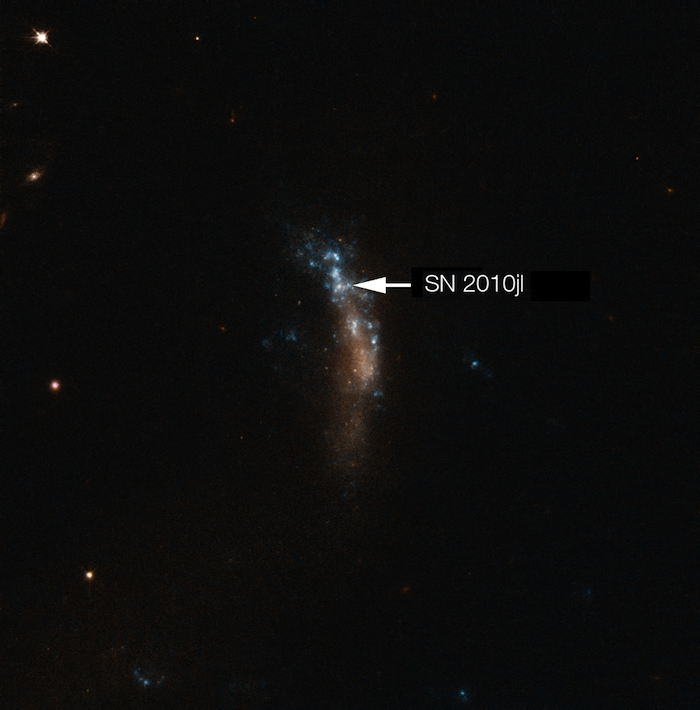In der Mitte des Bildes eine längliche Struktur die blau und orange leuchtet. Vereinzelt sind Punkte zu erkennen. Ein kleiner Punkt ist mit einem weißen Pfeil mit SN 2010jl bezeichnet. Der Hintergrund ist schwarz mit vereinzelten, leuchtenden Punkten.
