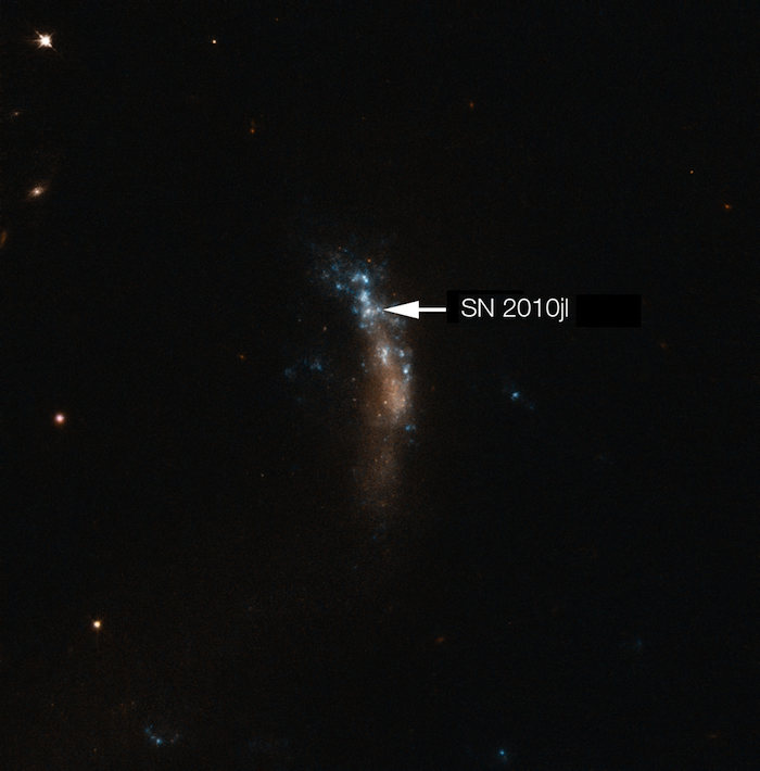 Supernova 2010jl In der Mitte des Bildes eine längliche Struktur die blau und orange leuchtet. Vereinzelt sind Punkte zu erkennen. Ein kleiner Punkt ist mit einem weißen Pfeil mit SN 2010jl bezeichnet. Der Hintergrund ist schwarz mit vereinzelten, leuchtenden Punkten.
