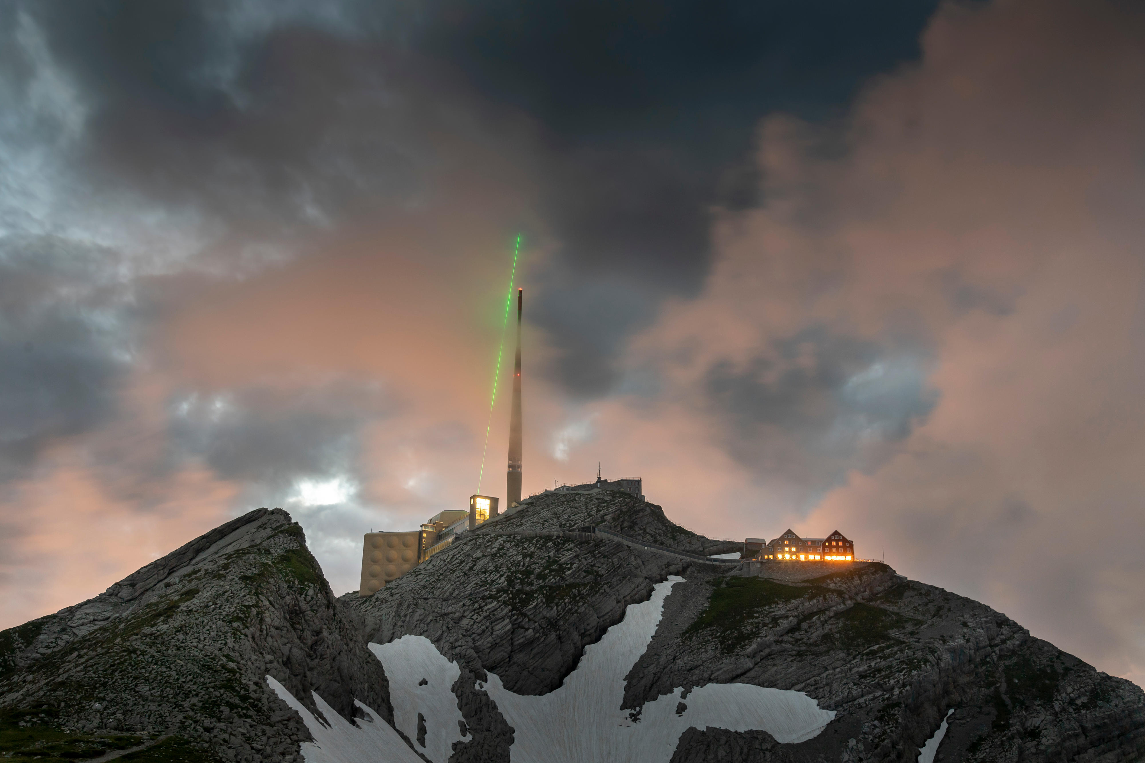 Auf einem Alpengipfel strahlt ein grüner Laser in einen verdunkelten Himmel über einem Antennenmast
