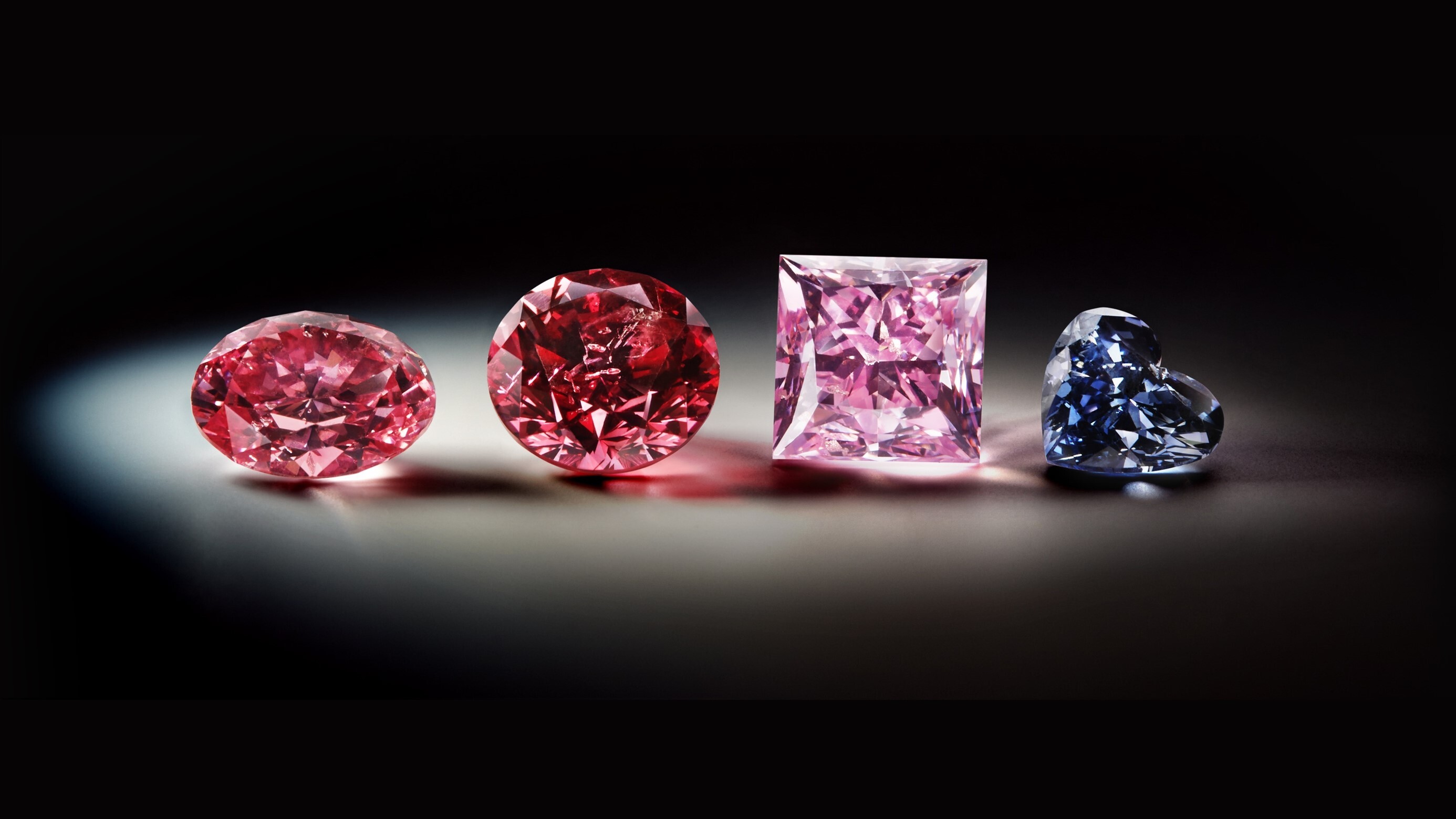 Vier verschieden geschliffene Diamanten vor schwarzem Hintergrund; drei davon sind rötlich gefärbt, einer blau
