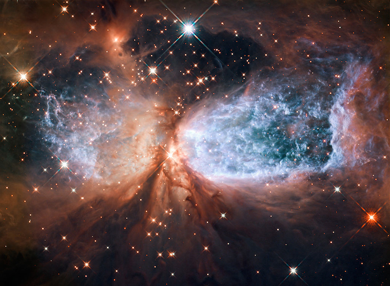 Sternentstehungsregion, aufgenommen vom Weltraumteleskop Hubble, Bildquelle: NASA/ESA; Lizenz: gemäß den Bedingungen der Quellen Eine Wolke aus rostrot leuchtendem Staub von dunklem Hintergrund mit einzelnen, funkelnden Sternen. Links und rechts vom Bildzentrum sind zwei runde Ausschnitte, in denen der Nebel blau leuchtet